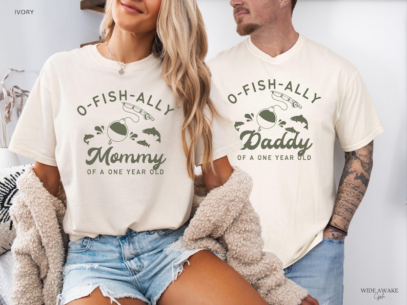 Pu&ograve; includere: T-shirt color avorio con grafiche coordinate. Una maglietta dice "Mommy of a One Year Old" e l'altra "Daddy of a One Year Old". Il design include un galleggiante da pesca, una canna da pesca e dei pesci.
