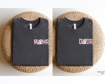 May include: Two dark gray t-shirts with the words "Mama" and "Dada" printed on the front. Each word is in a colorful, playful font with a cartoon character. The shirts are displayed on a woven, circular surface.