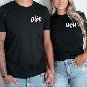 Puede incluir: Dos camisetas negras con dise&ntilde;os de manos de esqueleto blancas. La camiseta de la izquierda dice "DAD" y la camiseta de la derecha dice "MOM".