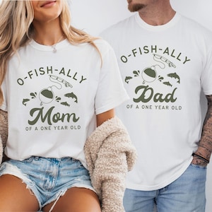 Pu&ograve; includere: T-shirt bianche con grafica a tema pesca. Una maglietta dice "O-FISH-ALLY Mom of a One Year Old" e l'altra dice "O-FISH-ALLY Dad of a One Year Old". Il design include un galleggiante, una canna da pesca e dei pesci.