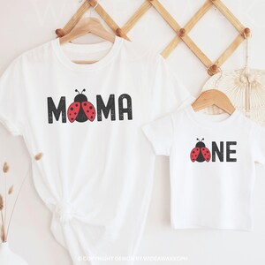 May include: Two white t-shirts, one adult and one child-sized, hanging on wooden hangers. The adult shirt says "MOMA" with a ladybug graphic. The child's shirt says "ONE" with a ladybug graphic. The shirts are displayed against a neutral background.