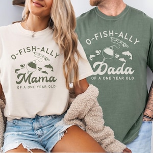 Pu&ograve; includere: Due magliette, una color avorio e una verde muschio, con grafiche a tema pesca abbinate. La maglietta avorio dice "O-Fish-ally Mama of a One Year Old". La maglietta verde muschio dice "O-Fish-ally Dada of a One Year Old".