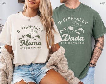Könnte beinhalten: Zwei T-Shirts, eines in Elfenbein und eines in Moosgrün, mit passenden Angelmotiven. Das elfenbeinfarbene Shirt trägt die Aufschrift "O-Fish-ally Mama of a One Year Old". Das moosgrüne Shirt trägt die Aufschrift "O-Fish-ally Dada of a One Year Old".