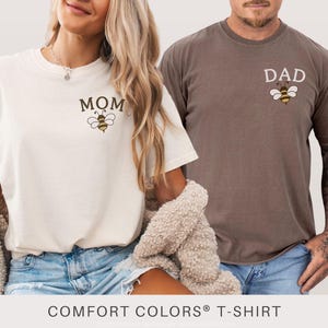 May include: Ivory and espresso Comfort Colors t-shirts. The ivory shirt has "MOM" and a bee graphic, while the espresso shirt has "DAD" and a bee graphic. Both shirts are short-sleeved.