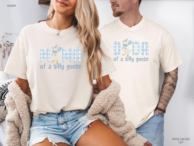 K&ouml;nnte beinhalten: Zwei elfenbeinfarbene T-Shirts mit einem blau-wei&szlig;en Vichy-Karomuster. Das linke T-Shirt zeigt den Schriftzug MAMA of a silly goose mit einer Gans. Das rechte T-Shirt zeigt den Schriftzug DADA of a silly goose mit einer Gans.