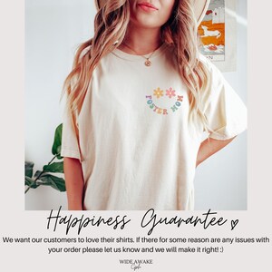 May include: A white t-shirt with a rainbow graphic that says "Foster Mom" with flowers around the text. The text "Happiness Guarantee" is written in black cursive with a heart symbol.