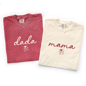 May include: Two t-shirts, one crimson and one ivory, are displayed. The crimson shirt has the word "dada" and a ladybug graphic. The ivory shirt has the word "mama" and a ladybug graphic. Each shirt has a silver necklace.