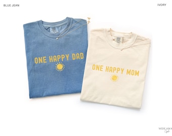 Op de afbeelding: Een blauw spijkerkleurig t-shirt met de gele tekst ONE HAPPY DAD en een klein zonnepictogram, en een ivoorkleurig t-shirt met de gele tekst ONE HAPPY MOM en een klein zonnepictogram. Beide shirts hebben een ronde hals en korte mouwen.