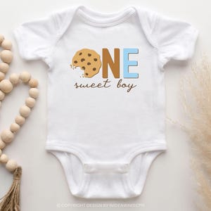 May include: White baby bodysuit with a brown and blue graphic that says "ONE sweet boy". The graphic features a chocolate chip cookie with a bite taken out of it.