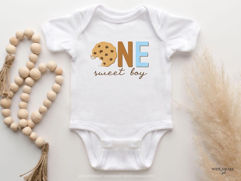 May include: White baby bodysuit with a brown and blue graphic that says "ONE sweet boy". The graphic features a chocolate chip cookie with a bite taken out of it.
