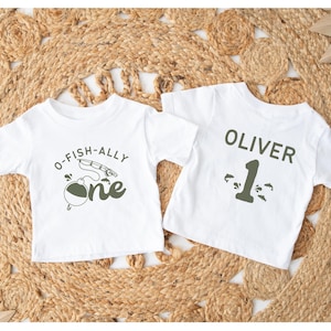 Pu&ograve; includere: Due magliette bianche per bambini piccoli. Una maglietta ha la scritta "O-FISH-ALLY one" con una grafica di canna da pesca. L'altra maglietta ha il nome "OLIVER" e il numero "1" stampati. Le magliette sono su una superficie beige intrecciata.