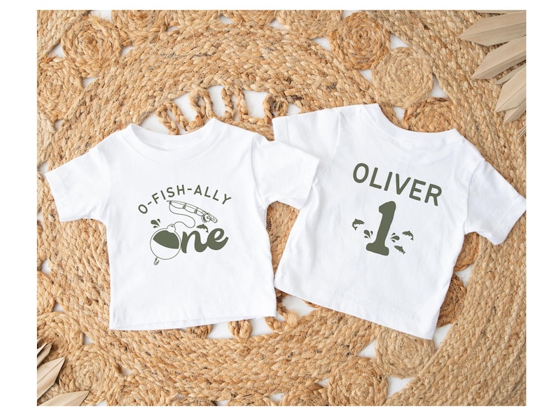 Pu&ograve; includere: Due magliette bianche per bambini piccoli. Una maglietta ha la scritta "O-FISH-ALLY one" con una grafica di canna da pesca. L'altra maglietta ha il nome "OLIVER" e il numero "1" stampati. Le magliette sono su una superficie beige intrecciata.