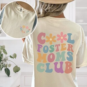 May include: A white t-shirt with the text "Cool Foster Moms Club" in colorful, retro-style letters with flowers. The back of the shirt has a smaller version of the text in a circle with flowers.