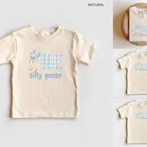 K&ouml;nnte beinhalten: Ein naturfarbenes Kurzarm-T-Shirt mit einer Grafik einer Gans, die einen blau gestreiften Partyhut und eine Fliege tr&auml;gt und einen blauen Luftballon h&auml;lt. Der Text lautet ONE in blauem Vichy-Muster und silly goose in blauer Schreibschrift.