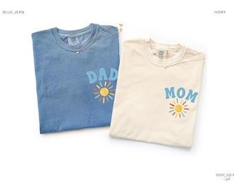 Op de afbeelding: Twee T-shirts met korte mouwen en een ronde hals, één in spijkerblauw en één in ivoor. Het blauwe shirt heeft de tekst DAD boven een zongrafiek, terwijl het ivoorkleurige shirt de tekst MOM boven een zongrafiek heeft. Beide shirts hebben een label.
