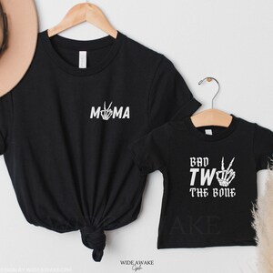 Puede incluir: Dos camisetas negras con texto blanco. La camiseta m&aacute;s grande dice "MOMA" con una mano de esqueleto haciendo un signo de paz. La camiseta m&aacute;s peque&ntilde;a dice "Bad Two The Bone" con una mano de esqueleto haciendo un signo de paz.
