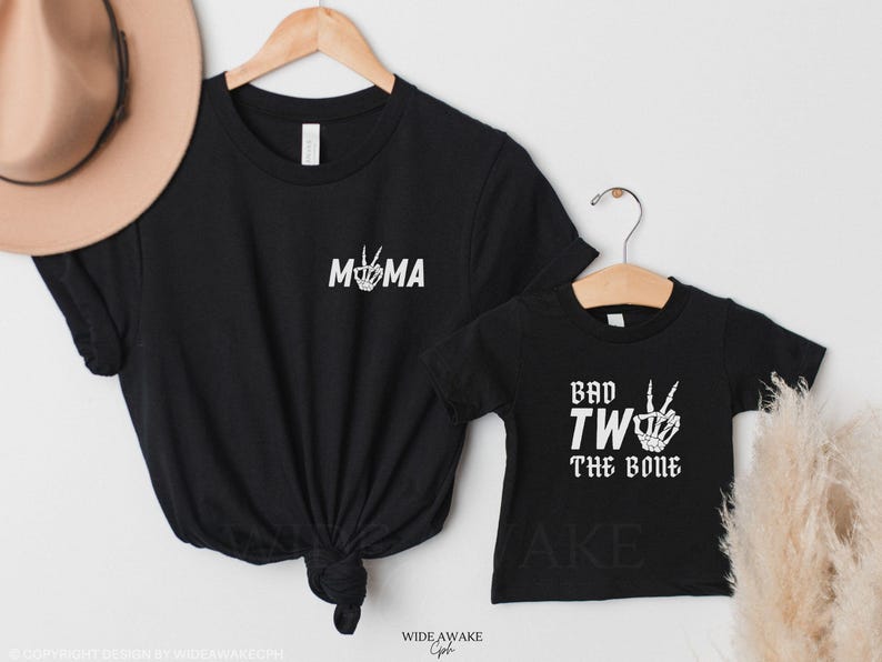 Puede incluir: Dos camisetas negras con texto blanco. La camiseta m&aacute;s grande dice "MOMA" con una mano de esqueleto haciendo un signo de paz. La camiseta m&aacute;s peque&ntilde;a dice "Bad Two The Bone" con una mano de esqueleto haciendo un signo de paz.