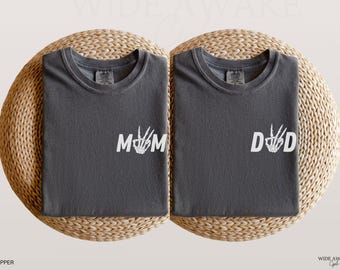 May include: Two charcoal gray t-shirts featuring white screen-printed graphics. The left shirt displays the text MOM with a skeleton hand forming the letter O. The right shirt displays the text DAD with a skeleton hand forming the letter A.