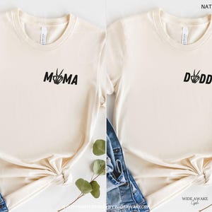Puede incluir: Dos camisetas de color natural con texto negro. La camiseta de la izquierda dice "MAMA" con un gesto de paz con la mano. La camiseta de la derecha dice "DADDY" con un gesto de paz con la mano.