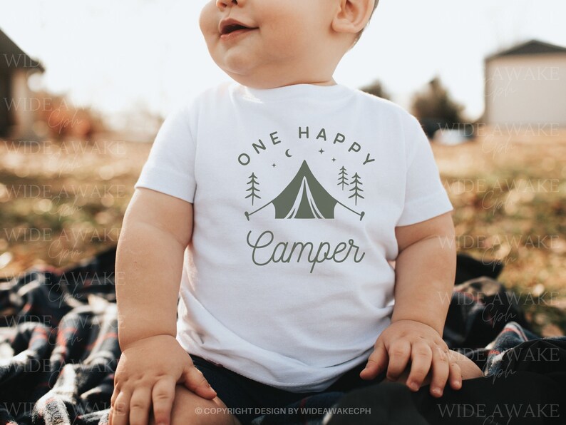Op de afbeelding: Een witte baby romper met een groene afbeelding van een tent, bomen en sterren. De tekst "One Happy Camper" is geschreven in een cursief schrift.
