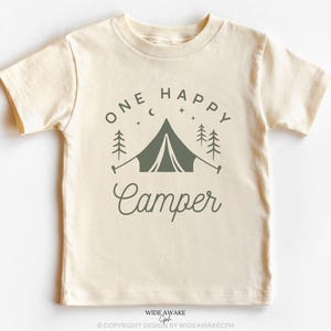 Op de afbeelding: Een cr&egrave;mekleurig T-shirt met korte mouwen met de tekst "One Happy Camper" in groene letters. Het ontwerp bevat een groene tent, bomen en sterren.