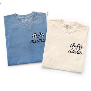 Könnte beinhalten: Zwei T-Shirts, eines in Blau und eines in Elfenbein, werden auf weißem Hintergrund gezeigt. Jedes Shirt hat ein gesticktes Design aus zwei karierten Flaggen und einem Wort. Das blaue Shirt sagt "mama" und das elfenbeinfarbene Shirt sagt "dada".