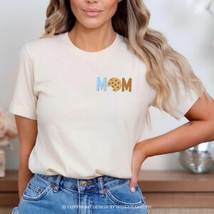 May include: A white t-shirt with the word "MOM" spelled out in blue and brown letters. The "O" is replaced with a chocolate chip cookie.