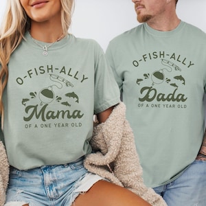 Pu&ograve; includere: Due magliette verde chiaro con la scritta "O-FISH-ALLY Mama" e "O-FISH-ALLY Dada" in verde scuro. Le magliette presentano grafiche a tema pesca, tra cui una canna da pesca, un galleggiante e dei pesci. &Egrave; incluso anche il testo "OF A ONE YEAR OLD".