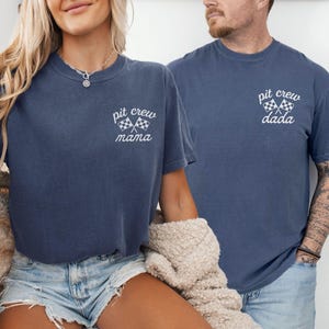 May include: Two denim-colored crew neck t-shirts featuring white graphic text and crossed checkered racing flags. The left shirt displays the text "pit crew mama" and the right shirt displays the text "pit crew dada" in a cursive and print font combination.