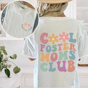 May include: White t-shirt with a colorful graphic that says "Cool Foster Moms Club" with flowers. The shirt also has a smaller graphic on the front that says "Foster Mom" with flowers.