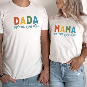 Peut inclure: Deux t-shirts blancs avec du texte coloré. L'un dit "DADA of the fun one" et l'autre "MAMA of the fun one". Le texte est orange, vert et bleu avec des étoiles. Les chemises sont portées par deux personnes.