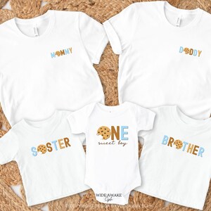 May include: A set of five white t-shirts with blue and brown cookie-themed designs. The shirts say "Mommy", "Daddy", "Sister", "Brother", and "One Sweet Boy".