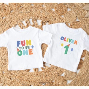 May include: Two white toddler t-shirts with colourful text. One says "FUN TO BE ONE" with a party hat and balloon. The other says "OLIVER" and "1" with balloons and a party hat. The shirts are on a woven mat.