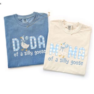 K&ouml;nnte beinhalten: Zwei kurz&auml;rmelige T-Shirts mit Rundhalsausschnitt in Jeansblau und Elfenbein. Beide zeigen eine Gans mit Partyhut und Schleife sowie den Schriftzug DIDA oder MIMA of a silly goose in blau-wei&szlig; karierten Blockbuchstaben im Vichy-Muster.