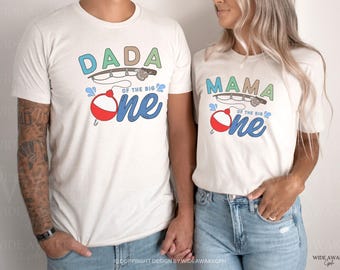 May include: Two white t-shirts with a fishing theme. The shirt on the left says "Dada of the Big One" with a fishing rod and bobber. The shirt on the right says "Mama of the Big One" with a fishing rod and bobber.