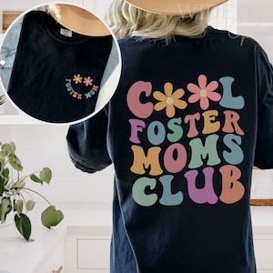 May include: Black t-shirt with a colorful graphic that says "Cool Foster Moms Club" with a flower design. The shirt has a small flower design on the front.