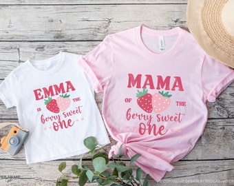 May include: Two white t-shirts with red text and a strawberry graphic. The larger shirt says "Mama of the berry sweet one" and the smaller shirt says "Emma is the berry sweet one".