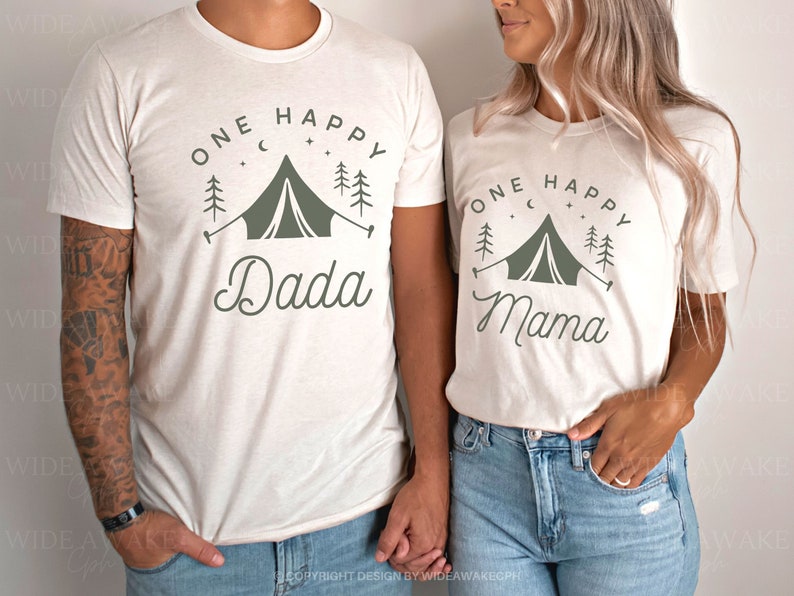 Op de afbeelding: Twee witte T-shirts met een kampeerthema. De shirts hebben een groene tent, bomen en sterren. E&eacute;n shirt zegt "One Happy Dada" en de andere zegt "One Happy Mama".