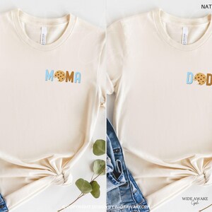 May include: Two natural colored t-shirts with a cookie graphic and the words "MOMA" and "DODA" in blue and brown letters.