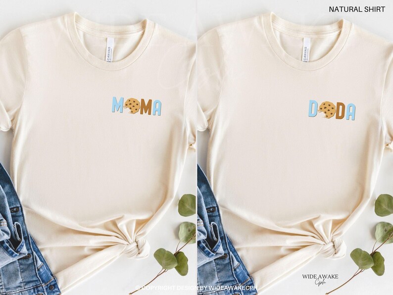 May include: Two natural colored t-shirts with a cookie graphic and the words "MOMA" and "DODA" in blue and brown letters.