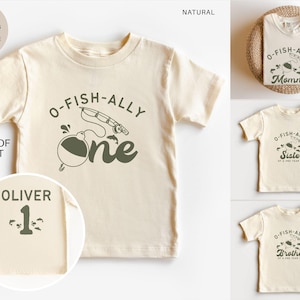 Pu&ograve; includere: T-shirt color crema a maniche corte con la scritta "O-Fish-Ally One" e una grafica a tema pesca. La parte posteriore della maglietta include il nome "Oliver" e il numero "1". Sono mostrate anche magliette aggiuntive con "Mommy", "Sister" e "Brother".