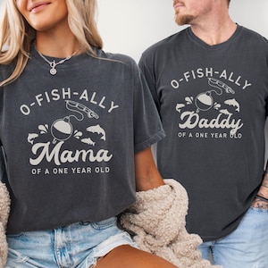 Pu&ograve; includere: Due magliette grigio scuro con la scritta "O-FISH-ALLY Mama of a One Year Old" e "O-FISH-ALLY Daddy of a One Year Old" in bianco. Le magliette presentano una grafica a tema pesca con un galleggiante, una canna da pesca e dei pesci.