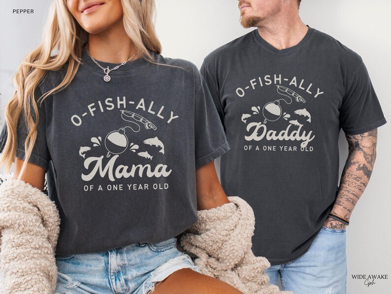 Pu&ograve; includere: Due magliette grigio scuro con la scritta "O-FISH-ALLY Mama of a One Year Old" e "O-FISH-ALLY Daddy of a One Year Old" in bianco. Le magliette presentano una grafica a tema pesca con un galleggiante, una canna da pesca e dei pesci.