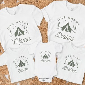 Op de afbeelding: Set van vier witte T-shirts met een kampeerthema. De shirts hebben een groene tent, bomen en sterren met de tekst "One Happy" en een ander woord op elk shirt: "Mama", "Daddy", "Camper" en "Brother".
