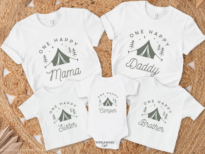 Op de afbeelding: Set van vier witte T-shirts met een kampeerthema. De shirts hebben een groene tent, bomen en sterren met de tekst "One Happy" en een ander woord op elk shirt: "Mama", "Daddy", "Camper" en "Brother".