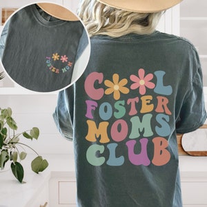 May include: A dark gray t-shirt with a colorful retro design that says "Cool Foster Moms Club" with flowers on the top. The shirt has a smaller design on the front that says "Foster Mom" with flowers.