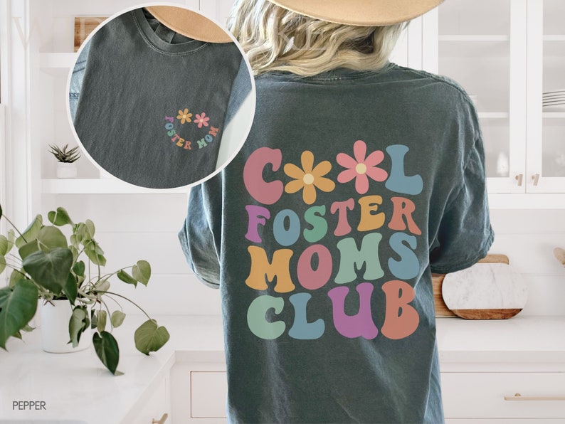 May include: A dark gray t-shirt with a colorful retro design that says "Cool Foster Moms Club" with flowers on the top. The shirt has a smaller design on the front that says "Foster Mom" with flowers.