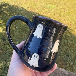 May include: A dark blue ceramic mug with a glossy finish, featuring a repeating pattern of small, speckled white ghost illustrations. The mug has a curved handle and a wide rim, showcasing a handcrafted appearance with visible texture.