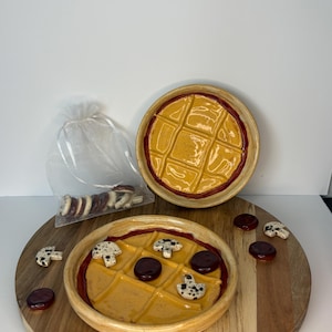 May include: Two round ceramic game boards with a grid pattern, featuring a glossy yellow surface and a dark red border. The boards are accompanied by small, speckled mushroom-shaped pieces and dark red circular tokens, with extra pieces in a bag.