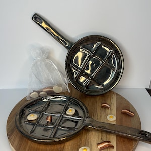 May include: Two dark metallic, circular ceramic pans with a grid pattern molded into the base. One pan is positioned upright, while the other lies flat. Small, realistic clay-like miniature fried eggs and bacon strips are scattered around.